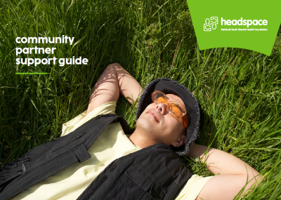 Cover of a headspace Community Partner Support Guide, featuring a young person wearing orange glasses, a black bucket hat and black vest, lying in tall green grass. White text reads “community partner support guide” with the headspace logo in the top right.