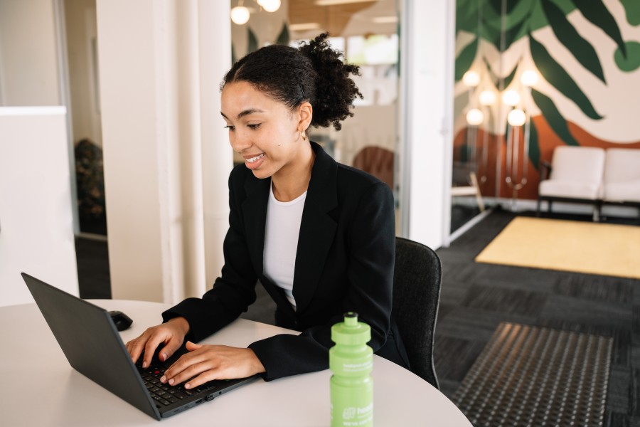 Person seated at a round table using a laptop in a modern office. A light green water bottle sits on the table in the foreground. The workspace features glass-walled rooms, decorative lighting, patterned carpet, and a large wall mural with green leaf designs in the background.