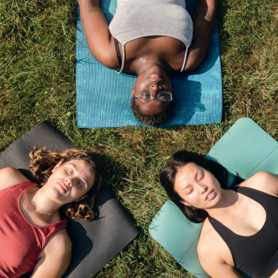 Overhead view of three people lying on yoga mats arranged in a triangle on a grassy field. Each person rests on a separate mat in different colours, with eyes closed and arms relaxed at their sides. Bright sunlight casts natural shadows across the grass, creating a peaceful outdoor wellness setting.