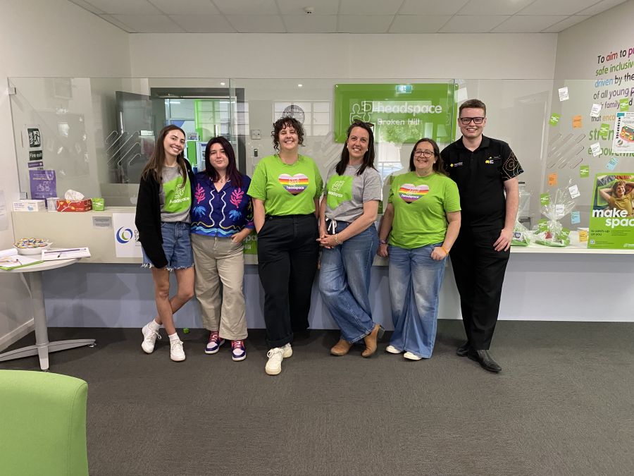 Six people standing together inside a Headspace centre reception area. Several people wear green Headspace-branded shirts, while others wear casual clothing. Behind the group is a reception counter with a green "headspace Broken Hill" sign, informational displays, posters, and notes on the wall. The space features glass partitions, grey carpet, and bright indoor lighting.