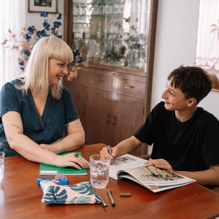 Two people sit at a wooden dining table in a home setting, working together on schoolwork. One person is writing in an open workbook while the other points to a notebook on the table. Glasses of water, pens, a pencil case and study materials are spread across the table. In the background, sheer curtains, a display cabinet and framed wall art are visible.
