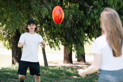 A person in a light-coloured T-shirt, black shorts and a black cap stands on grass in a park, reaching toward a red Australian rules football suspended in mid-air. Another person is visible in the foreground with their back to the camera. Tall leafy trees fill the background, and the scene is lit by daylight.