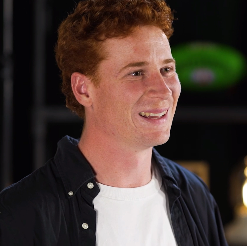 Close-up portrait of a person with curly red hair wearing a white T-shirt and a dark button-up shirt. The person is facing slightly to the side indoors, with a blurred background featuring dark shelving and soft lighting. No visible text appears in the image.