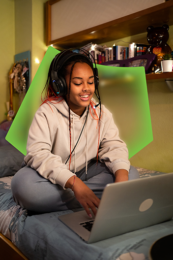A young person sitting on their bed with headphones on, looking at their laptop. The young person is smiling and appears to be relaxed.