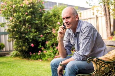 An older man sitting in his garden on a bench speaking on his mobile phone.