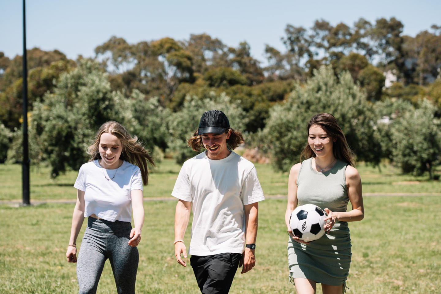 Three young people in a park, walking towards the camera, with one holding a soccer ball.