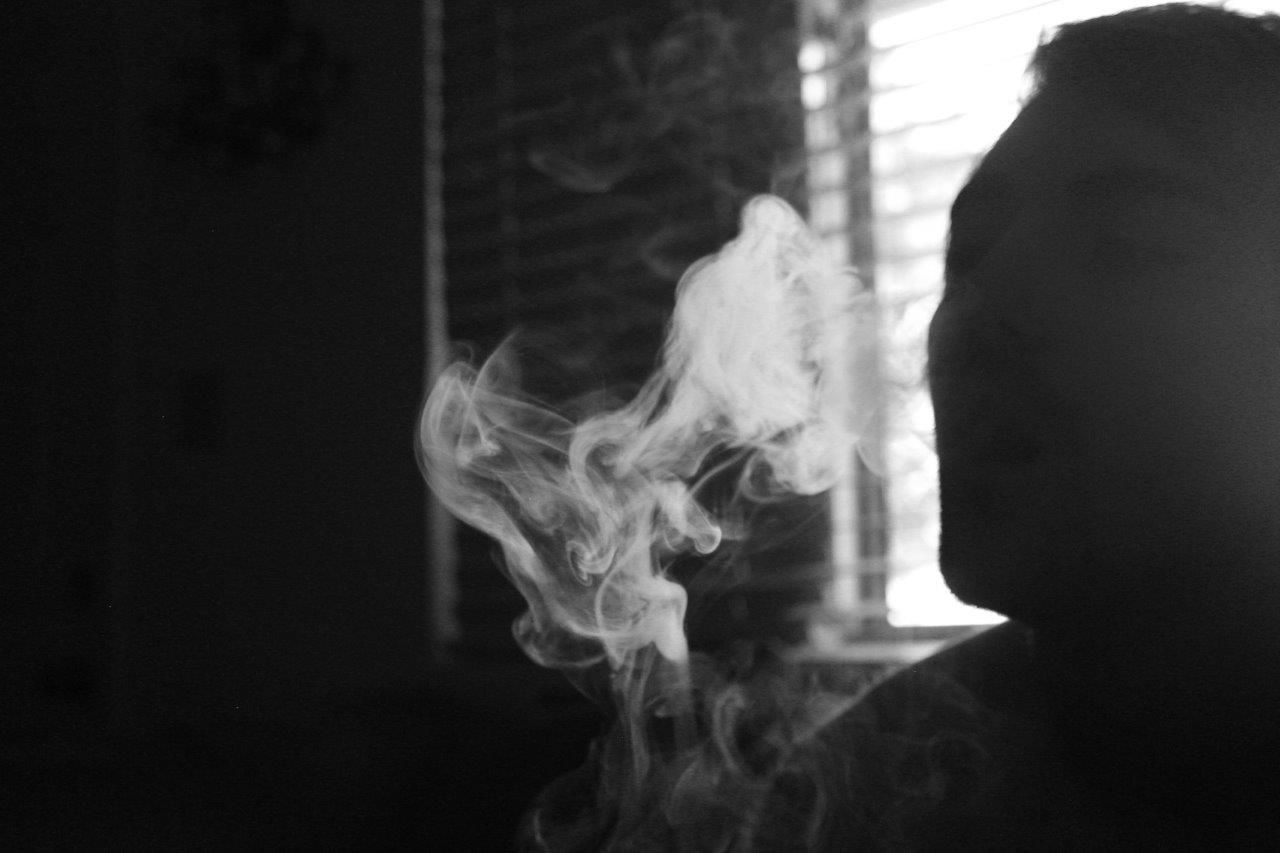 Almost silhouetted face blows smoke out in a dimly lit room, black and white.