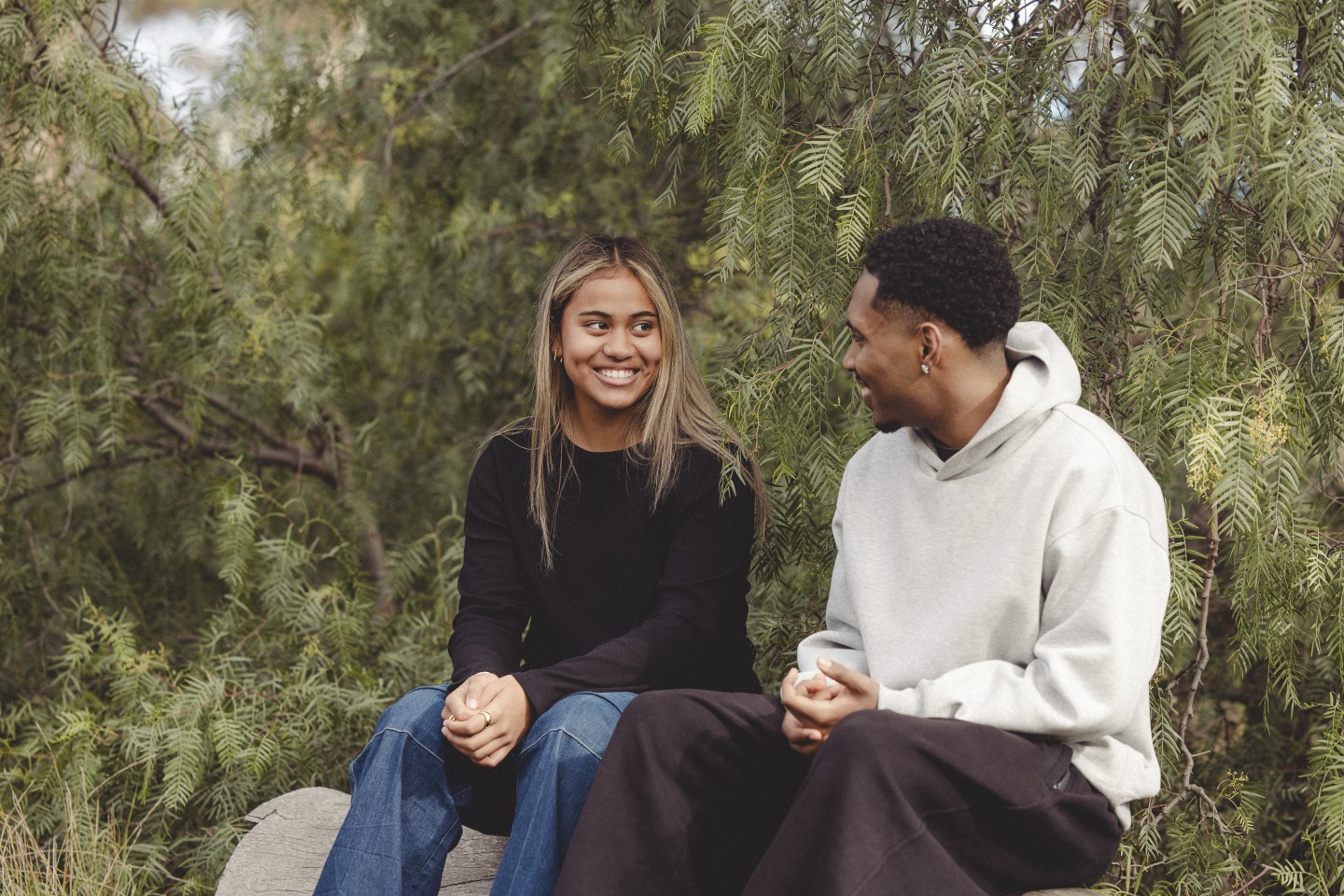 Two young people sit outdoors on a bench surrounded by greenery, smiling and talking with each other in a relaxed, friendly moment.