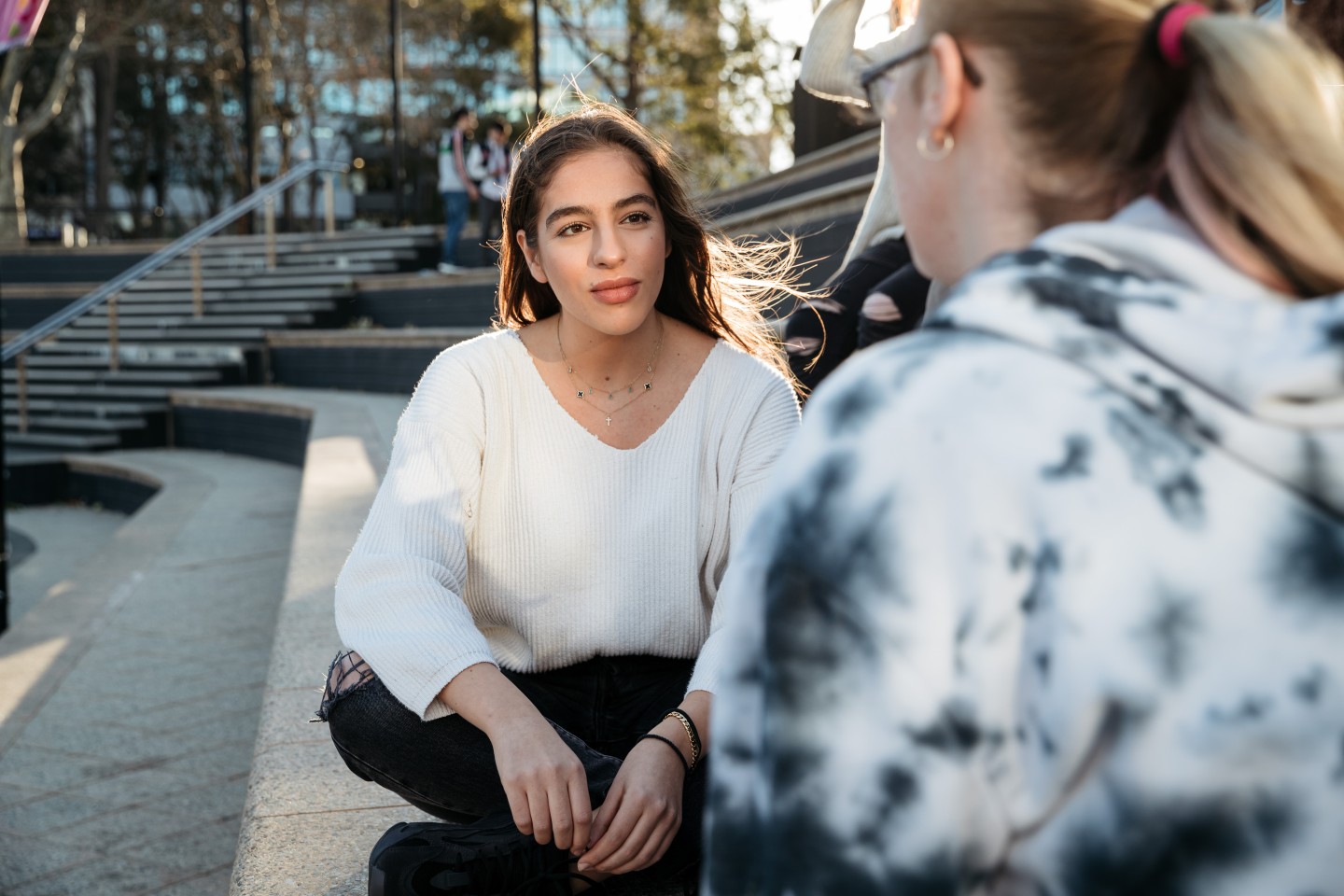 Two people are engaged in a conversation outdoors. One person, wearing a white sweater and black pants, is sitting on the steps and facing the other person. The second person, whose back is to the camera, is wearing glasses and a tie-dye hoodie. The background shows more people and steps leading up to an area with trees and buildings
