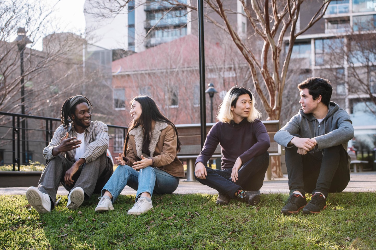 Four people sitting on grass in an urban park, engaged in conversation. Leafless trees, buildings, and a bench are visible in the background