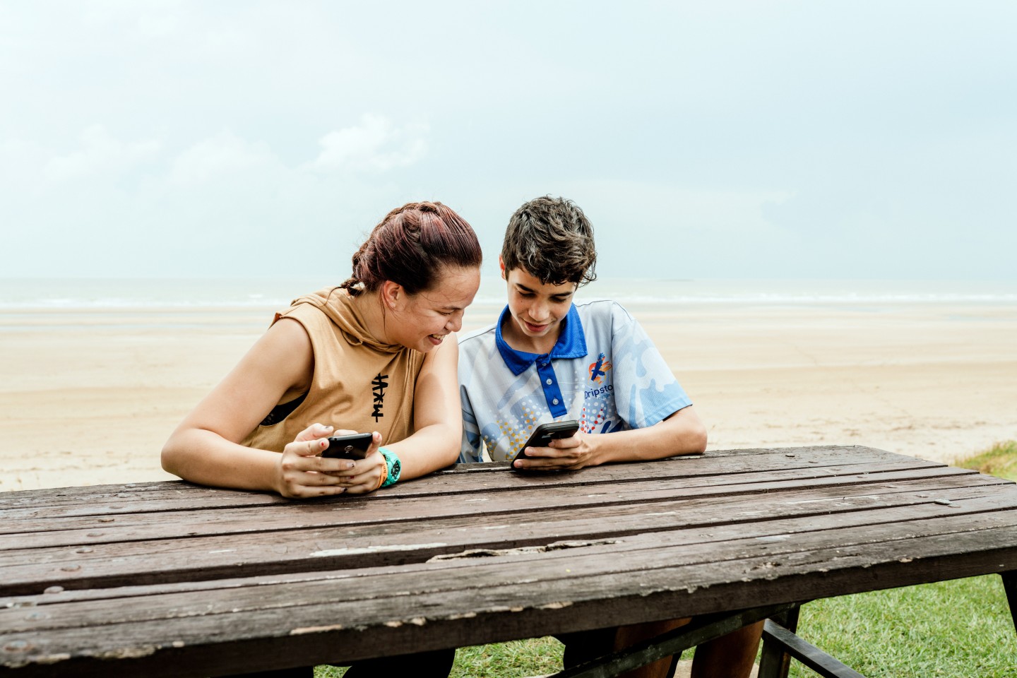 A teenage girl and a teenage boy sit at a wooden picnic table by the beach, smiling and looking at their phones together. The beach and ocean are in the background under a cloudy sky.