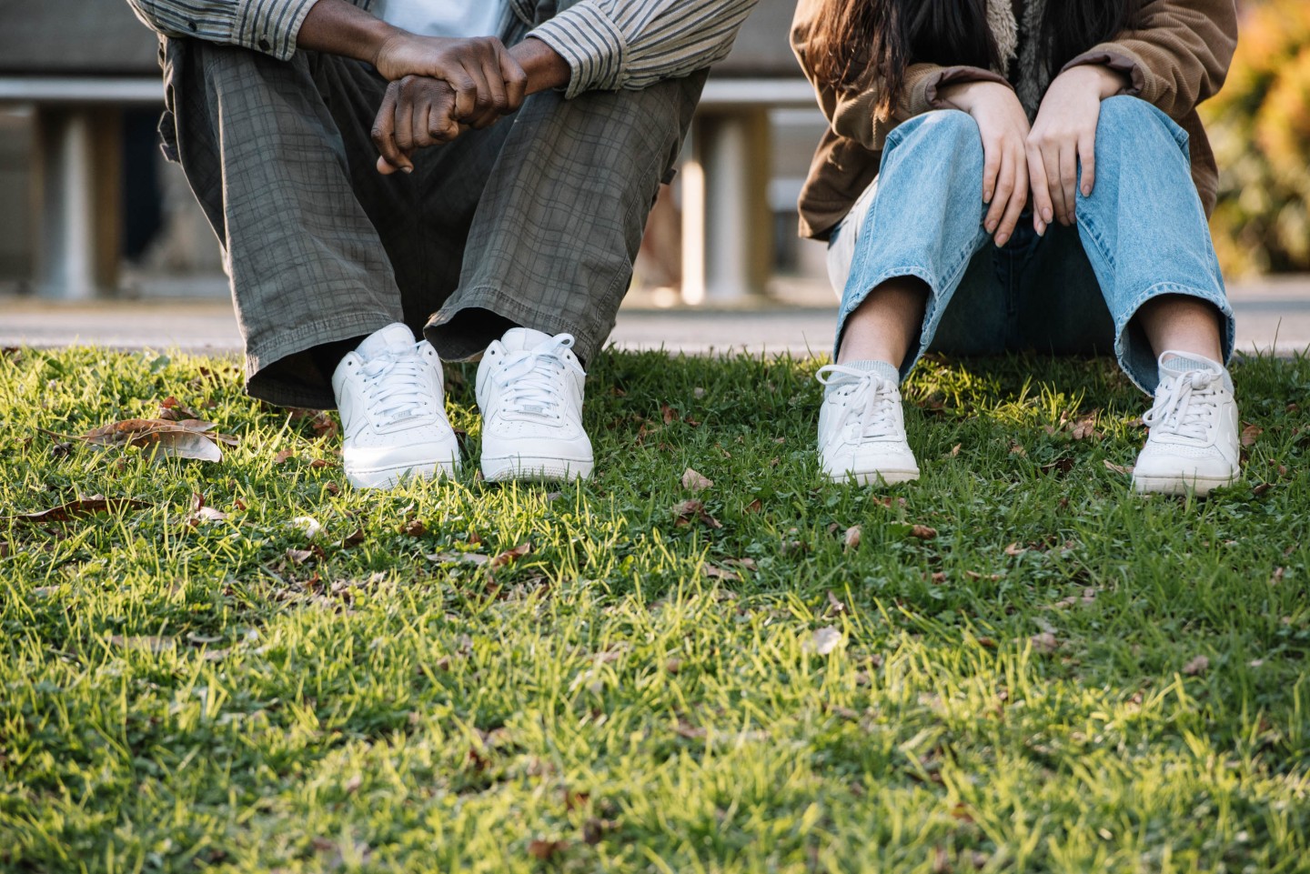 Two young people sitting side by side on grass outdoors, with only their lower bodies visible. One person rests their hands together while the other sits with knees pulled up. Both are wearing casual clothes and white sneakers in a calm outdoor setting.