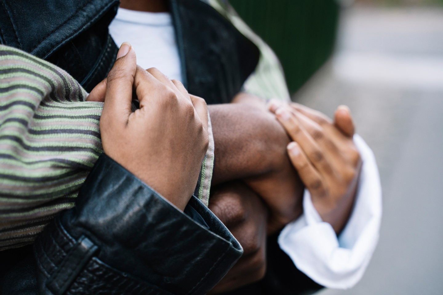 Close-up of two people embracing, with one person gently holding the other’s arm. The image focuses on their hands and layered clothing, including a striped sleeve and black leather jacket, conveying comfort, support and connection.