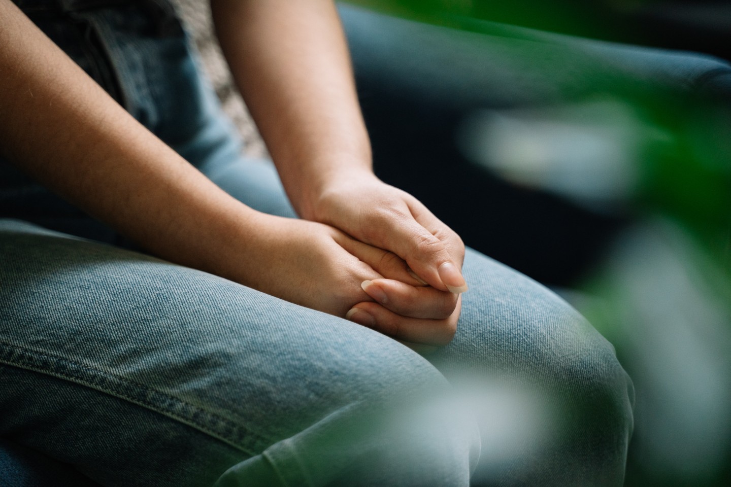"Close-up of a person sitting with their hands clasped together on their lap, wearing light blue jeans."