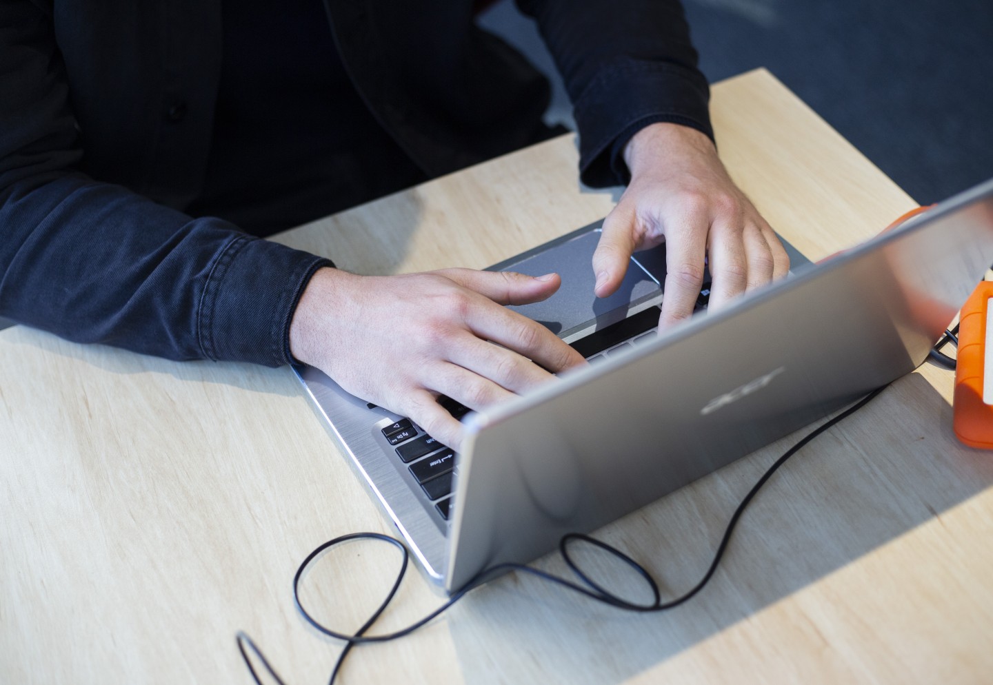 "Close-up of a person's hands typing on a silver Acer laptop, placed on a light wooden desk. A black cable is plugged into the laptop, and an orange external hard drive is visible on the right side."