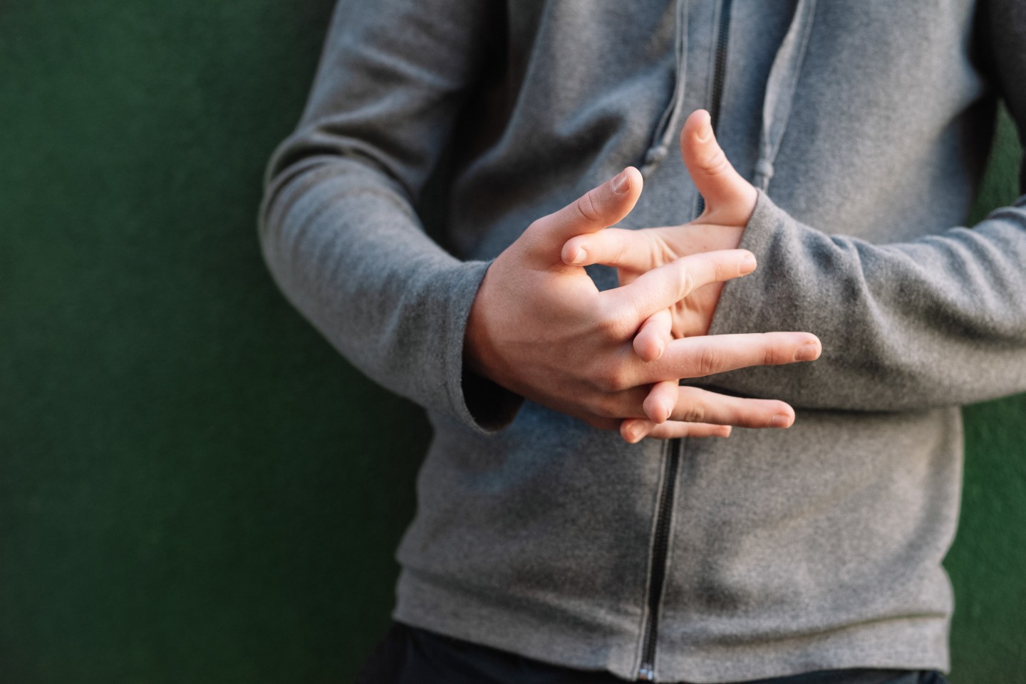 Close-up of a person wearing a grey zip-up hoodie, standing against a dark green background with their fingers interlaced and hands fidgeting in front of their body.