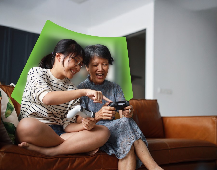 A mother and daughter sitting on a couch. They are both smiling. The mother is holding a video game controller and the daughter is pointing to a button, teaching the mother how to use the controller. 