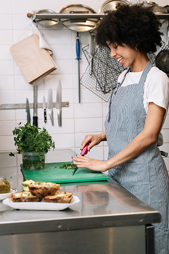 A young person standing in a commercial kitchen at a bench. The young person is wearing a blue apron and is chopping fresh Parsley leaves.