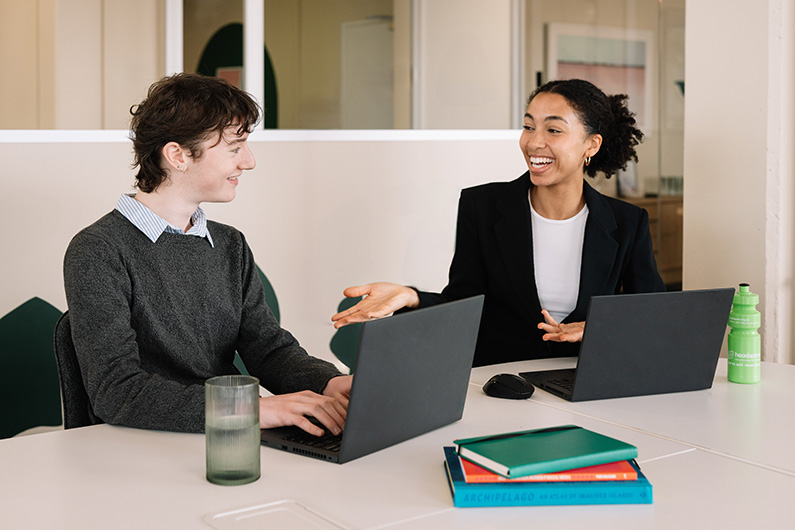 Two young people sitting at a table with their laptops, talking to each other happily. happ 