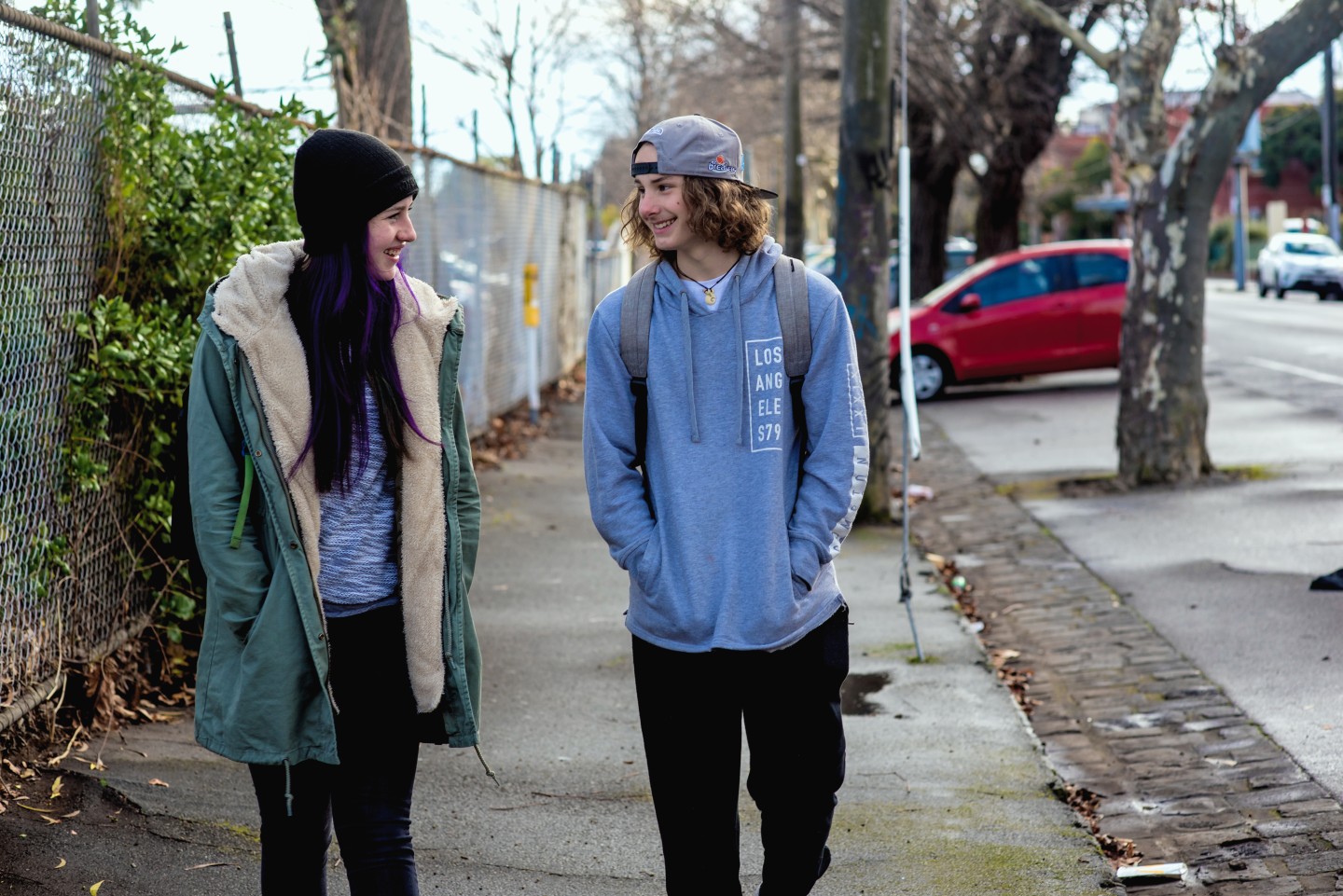 Two young people talking to each other as they walk down an urban street. One a beanie and a long coat, the other wearing a backwards cap and a hoodie.