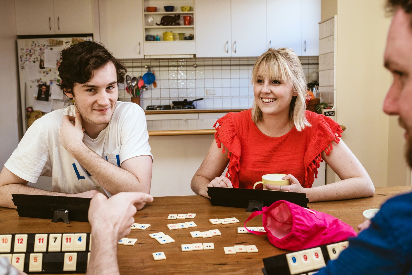 Family playing a board game at a dining table, with two young people facing the camera, and a man to the right is slightly out of frame.